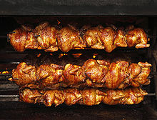 Six golden-brown whole chickens are being roasted on each of three black metal spits.