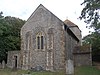 A flint rubble church with large stone quoins. In the nearest wall is a two-light lancet window between buttresses. A squat tower with a squared-off pyramidal roof is partly obscured behind this.