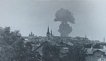 Schwarzweißfotografie von einer Stadt mit einer riesigen Rauchwolke. Einige Kirchtürme ragen hervor und vorne bedecken Bäume manche Häuser.