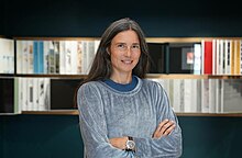 Manuela Pfrunder Smiling, arms crossed, wiht a soft, blurred background of bookshelves