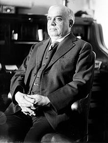 Black and white photo of a heavy elderly gentleman, seated, wearing a dark three piece suit