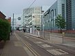 A paved road with one tram track in the middle and a tram shelter on the right side. There is a glass facade on the outside of a shopping centre in the background.