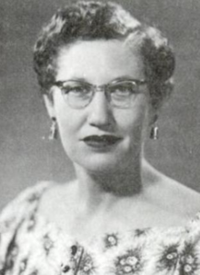 A white woman with short dark hair parted at the side, wearing glasses, earrings, dark lipstick, and a print dress with a wide square neckline