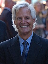 A headshot of a smiling man with long white hair and a black suit