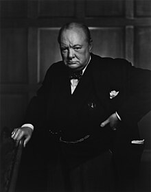 Churchill, 67, wearing a suit, standing and holding into the back of a chair