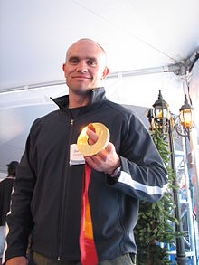 A hair-shaved man smiles at the camera while holding in his left hand a golden medal. He wears a dark sports jacket with a white stripe along the arm length, and is inside a large reception tent with a small street lamp and a pine tree behind.