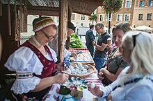 Na zdjęciu stragan podczas Jarmarku Jagiellońskiego. Za ladą kobieta ubrana w strój ludowy, sprzedaje pisanki. Przy stoisku stoją klienci.