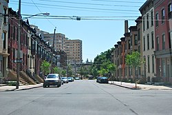 A view down a city street from an intersection. On either side are rows of two-story brick houses, largely identical with their neighbors. In the background a taller building is visible to the left.