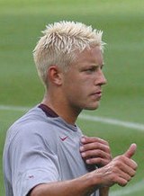 A photograph of a man with short blonde hair wearing a grey shirt with a maroon collar. He is giving someone the thumbs-up signal.