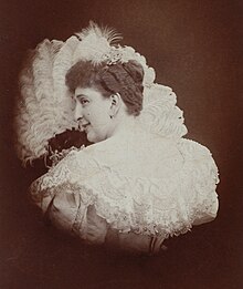 A white woman with curled hair, wearing a lace-trimmed white dress, posed in front of a feathery or ruffled fan