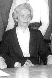 An older white woman, seated, with grey hair in a set style, wearing a white blouse and a dark cardigan