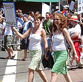 Zu sehen sind zwei Frauen die auf einer Straße in einer Menschenmenge Händchen halten. Beide tragen Röcke und ärmellose weiße Oberteile. Eine der Frauen links im Bild hat kurzes Haar, die andere langes. Die Kurzhaarige hält ein Schild hoch uf dem schlecht erkennbar die Worte "Married..." zu lesen sind.