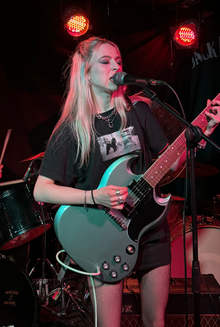 Full-body shot of Lizzie Esau, a young white, woman with bleached hair, wearing a dark T-shirt, shorts and fishnets, singing and playing an electric guitar.