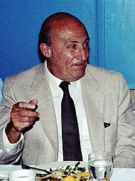 An bald elderly man in a suit and tie, seated before a meal, raising his right hand slightly and look to the right of the picture.