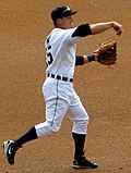 Baseball player in white uniform in a baseball throw followthrough position. His arm is forward with his hand down and his left hand is gloved. His left foot is planted and his right foot is in the air behind him.