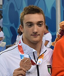 Burdisso trägt eine weiße Trainingsjacke mit dem italienischen Wappen, mit der rechten Hand zeigt er seine Bronzemedaille. Am rechten Bildrand ist der rote Trainingsanzug des russischen Siegers zu erkennen.