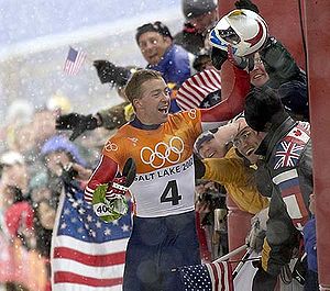 A man celebrates near a joyful crowd waving American flags behind a security fence. Holding a helmet high in his left hand, he wears a blue, red, white, and orange jumpsuit with the Olympic rings, the words Salt Lake City, and a number four in the chest.