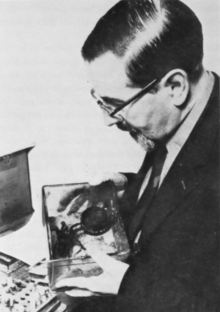 A man wearing glasses, a jacket, and tie is holding a clear box with a spider inside.