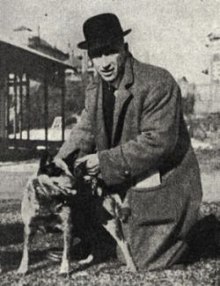 Ein Mann von etwa 30 Jahren mit dunklem Mantel und einem schwarzen Hut, einer Melone, auf dem Kopf. Er kniet auf dem Boden und streichelt einen mittelgroßen Hund