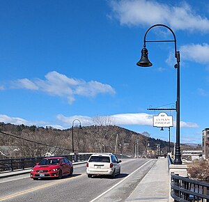 A photograph of the road deck of the Lyman Bridge, facing west, including a sign bearing its name, two vehicles, and hills in Vermont in the distance.