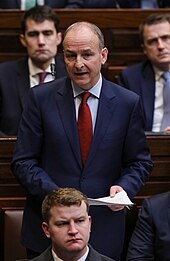 A man in a blue suit stands in a government debate chamber