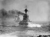 A large gray battleship steams in choppy seas; thick black smoke billows from its funnels. Two battleships are directly behind