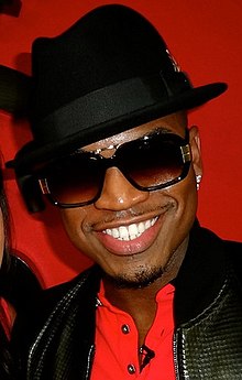 Ne-Yo steht vor einer roten Wand. Er trägt eine schwarze Lederjacke, ein rotes Hemd mit einem schwarzen Mini-Mikrofon an der linken Knopfleiste, eine eckige schwarze Sonnenbrille, einen kleinen Diamant-Ohrstecker im linken Ohr und einen schwarzen Fedora. Er trägt zudem einen dünnen Schnurr- und einen leichten Kinnbart.