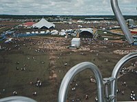 From a place up high and distant is viewed a vast open field of grass, trodden bare and brown in places with little people dotted around like ants beside enormous tents, marquees, performance stages hosting deafening arrays of musical-event speakers. The horizon is low and far away and the sky is dotted with broken clouds.