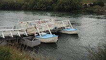 A dock spanning to a punt ferry in a river, a platform at the end of the dock resting on two small boats