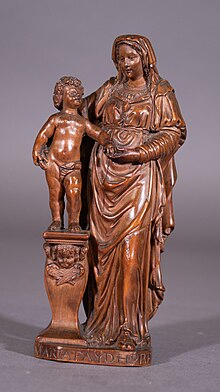 eine kleine rötlich-braune Buchsbaumfigur einer Madonna mit Kind. Das nackte Jesuskind steht auf einem Podest und reicht Maria bis zur Schulter. Am Fuß ist eine Signatur zu erkennen