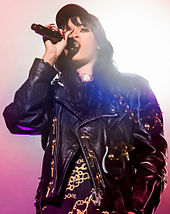Siyah bir şapka takmış olan siyah saçlı, siyah ceketli bir kadın mikrofon ile şarkı söylüyor