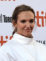 A middle-aged White woman with long, tied-back hair and wearing a white top stands in front of a white background with text.