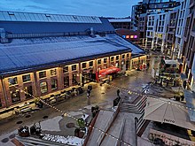 Foto auf einen Weg hinab, auf dessen rechten Seite sich höhere moderne Wohngebäude befinden. Links vom Weg befindet sich eine Gebäude mit roter Ziegelfassade.