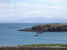 A two-masted yacht and a RIB lie at anchor on a sunny day off a grassy shore, with low cliffs beyond. A skerry lies further offshore to the left with high hills in the distance.