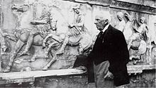Black-and-white photograph of an elderly man in a dark suit, in front of a wall of Ancient Greek sculptures.