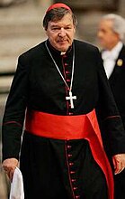 Kardinal George Pell memakai pakaian biasa kardinal: jubah hitam dengan perhiasan kain jalur merah, dan butang merah, fascia (selempang) merah, salib dada pada rantai, dan zucchetto merah.