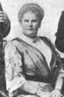 A white woman with light-colored bouffant hair, seated, wearing a loose satiny robe-like dress over a white high-collared blouse