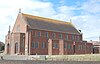 Three-quarter view of a tall, plain red brick church with an entrance porch at the left end and several uneven projections from the near side. The porch has a stone statue and an arched entrance door. The tiled roof has stone ends, both of which have small stone crosses. The lower level of the longer side has 15 unevenly spaced but identical round-headed windows; above this and immediately below the roofline are another eight windows of the same type, which are evenly spaced.