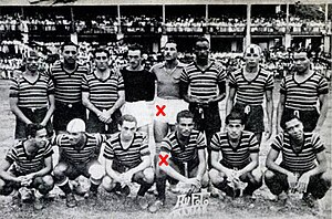 Fourteen football players, eight standing and six squatting. The fifth standing and the fourth squatting players from the left have been marked with a red cross.