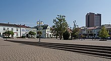 Zdjęcie rynku z perspektywy pieszego. Na pierwszym planie charakterystyczna, czerwona kostka. Dookoła pastelowe budynki stylizowane na stare kamienice i nieliczne drzewa. W tle wysoki budynek sądu z charakterystyczną, kanciastą bryłą.