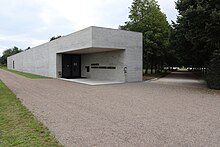 Ein flaches Gebäude aus Glas und Beton steht an der Ecke eines Feld- und eines Alleeweges