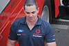 A man in his early thirties walking in a paddock. He is looking down from the camera and is wearing a grey T-shirt with sponsors logos.