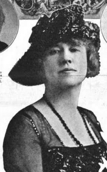 A white woman wearing a dark hat with an asymmetrical brim, and a dark dress with a square neck and translucent sleeves. She is also wearing dark beads.