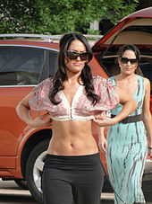 Identical female twins with long black hair are both walking towards the camera, one slightly in front of the other. The one in front is wearing a patterned, pastel colored crop top with black trousers, and is wearing her hair loose. The one behind is wearing a blue dress and has her hair tied back. Both are wearing sunglasses.