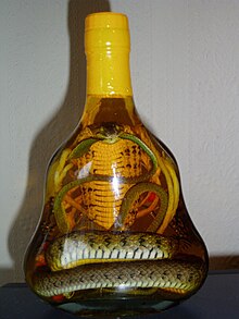 Eine Schlange steckt aufrecht in einer kegelförmigen Glasflasche mit gelbbrauner Flüssigkeit. Durch das Maul der Schlange läuft eine weitere dünne, grünliche Schlange. Der Hals ist breit wie bei einer Kobra.