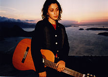 The flash of the camera lights up a woman holding an acoustic guitar with the sun setting on a body of water behind her.