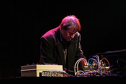 Carl Michael von Hausswolff standing on a darkened stage, bending over a set of electronic consoles on a table