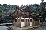 Small wooden building with white walls and hip-and-gable roof connected to another wooden structure in the back.