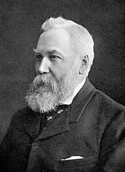 A head and shoulders view of an elderly man with white hair and a large, bushy beard. He is wearing a dark jacket and tie and a white shirt