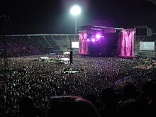 A stadium filled with spectators on the podiums and on the ground. In the middle is a stage with two giant pink 'L’ symbols flanking it. A large flood light is visible behind it.